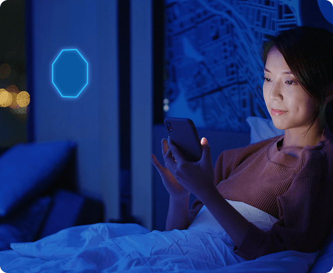 Woman relaxing in a hotel bed at night with her smartphone in hand. There is a glowing ADT emblem next to her, indicating the new My Safety service in the ADT+ app.